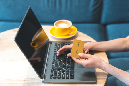 Cropped photo of adult and confident woman sitting in cafe with modern interior with soft and natural daylight. Female using her laptop and holding credit card in handの写真素材