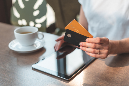 Close up cropped photo of young or adult woman sitting in cozy cafe with modern interior with soft and natural daylight. Female holding two credit card in hands and using tabletの写真素材