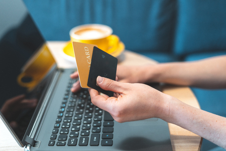 Cropped photo of adult girl or young woman spending weekend day in cozy cafe with comfort and modern interior with bright light room. Female using two bank card with computer to making online shoppingの写真素材