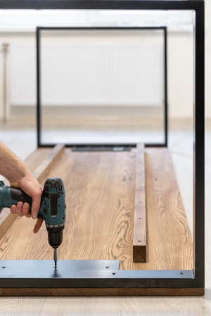 Vertical photo of professional handicraft with professional electric screwdriver instrument in hand and making repair or assemble new wooden desk or table top with metallic elementの写真素材