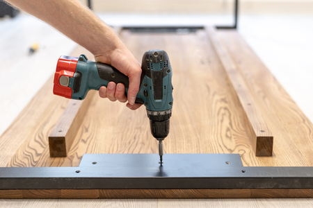 Side view and close up photo of carpenter person with professional electric screwdriver instrument in hand and repairing, fixing on building new wooden desk or table top with metallic legの写真素材