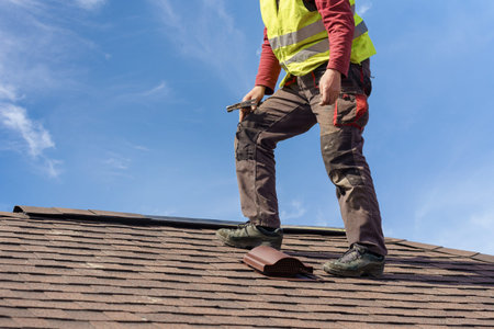 Unrecognizable workman standing on tile roof of new home under construction against blue sky with...