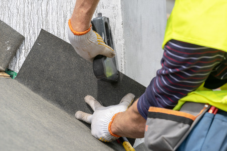 Close up, cropped photo hands with stapler of qualified and mature contractor in protective uniform wear. Man attach special waterproof bitumen membrane on roof top of new modern building constructionの写真素材