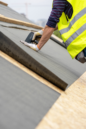Vertical, cropped and close up photo hands of mature and professional worker with stapler in hands working on rooftop of new modern building construction and attach special waterproof bitumen membraneの写真素材