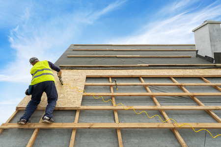 Concept of renovation, repair frame work house. Man holding pneumatic nail gun and fixing wooden roof cover. Qualified and mature contractor standing on rooftop of new modern building constructionの写真素材