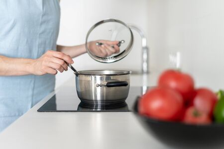 Cropped view of woman in blue apron holding spoon in hands, cooking food in saucepan, using built in electric stove, standing in white modern kitchen with bowl of vegetables at tableの写真素材