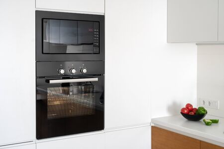 Black and modern built in oven with closed glass door on white contemporary kitchen with vegetables in plate on tableの写真素材