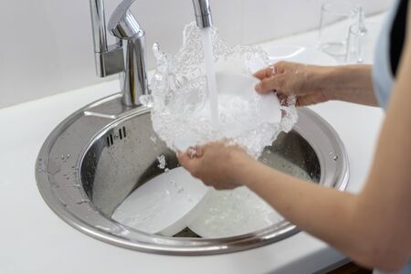 Cropped view of woman holding dishware in hands, washing plate in sink with water splash, standing on white bright kitchenの写真素材