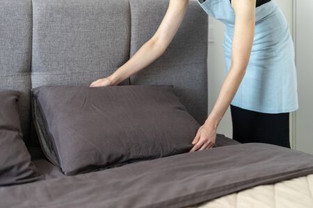 Cropped view of housemaid woman making bed, holding pillow in hands, standing inside bright and light bedroomの写真素材