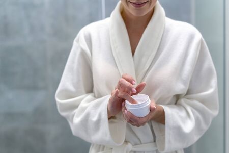 Cropped view of young adult woman in bathrobe holding jar with cream in hands, standing at bathroom with copy spaceの写真素材