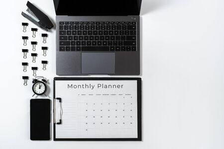 Top view of monthly planner in clipboard, laptop, smartphone, alarm clock, paperclip and stationery isolated on white background with copy spaceの写真素材