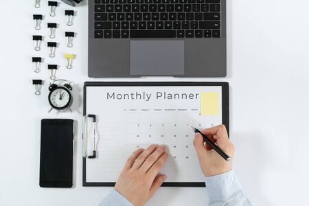 Top view of woman making notes in monthly planner. Laptop, smartphone, alarm clock, paperclip and stationery isolated on white background with copy spaceの写真素材
