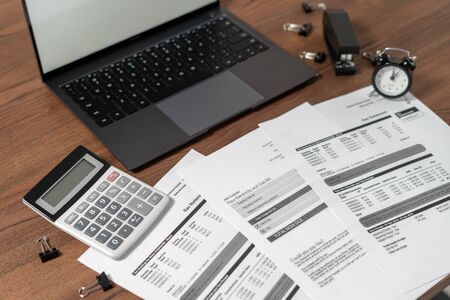 Modern laptop computer, documents, financial bills, alarm clock, calculator and stationery on wooden table in officeの写真素材