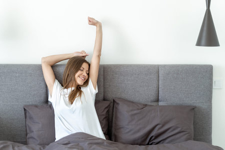 Young adult girl smiling, waking up in morning, stretching, raising hands up and sitting on comfort...