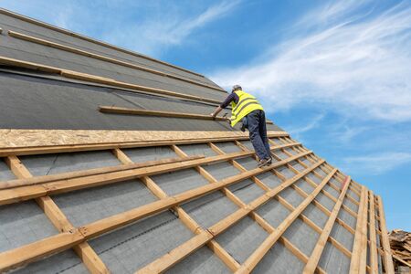 Concept of renovation, repair and fixing frame work house. Man hammer nails on wooden plank on rooftop. Unrecognizable mature worker standing on roof top of new modern building constructionの写真素材