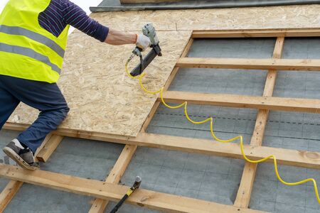 Concept of building construction industry. Cropped, unrecognizable, roffer or workman in protective uniform wear using air or pneumatic nail gun recovering roof top on new modern houseの写真素材