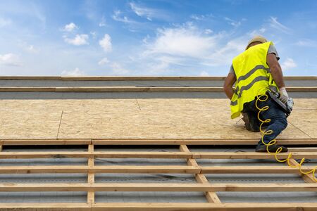 Concept of house under construction. Unrecognizable and professional workmen working on rooftop of new modern building. Man holding pneumatic nail gun in hands standing against blue sky on backgroundの写真素材