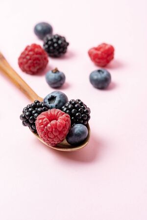 Vertical photo and close up view of wooden spoon with mix ripe and sweet blueberries, blackberries, raspberries fruits on pastel pink background with copy spaceの写真素材