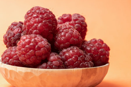 Macro view of red and ripe raspberry fruits in wooden bowl on pastel peach background with copy...