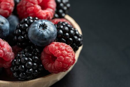 Close up view and macro of mix different ripe and sweet blueberries, blackberries, raspberries fruits in wooden bowl on black background with copy spaceの写真素材