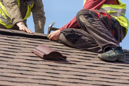 Two mature roofers in protective uniform using helmet and installing asphalt shingle or roof tile on top of new house against blue skyの写真素材