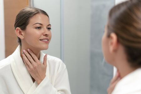 Young adult and good-looking woman in white bathrobe standing in bright light bathroom with mirror. She looking at her reflection, holding hand on neck and smilingの写真素材