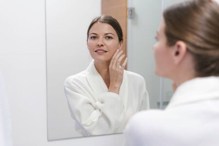 Young adult and good-looking woman in white bathrobe standing in bright light bathroom with mirror. She looking at camera and smiling cheerfulの写真素材