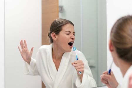 Funky young woman in white bathrobe standing in bathroom, holding tooth brush, looking at mirror and singing at toothbrush like in microphoneの写真素材