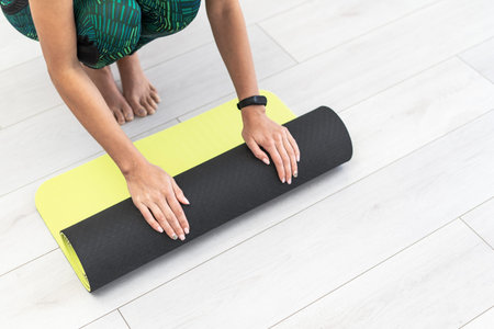 High angle view of young adult woman rolling fitness mat on the floor and preparing to training at...