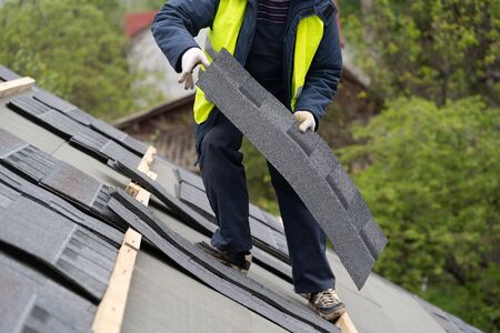 Qualified collar worker or roofer in protective uniform installing asphalt or bitumen tile on top of the roof under construction houseの写真素材