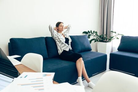 Side view of young adult woman sitting on sofa with closed eyes, holding hands behind head and relaxing at home after workの写真素材