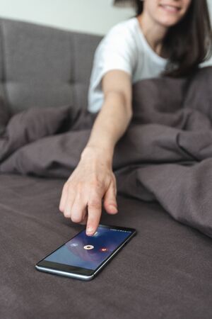 Selective focus of happy young adult woman waking up in bed and switch off alarm clock notification on modern smartphoneの写真素材