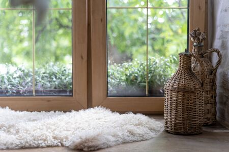 Comfort and cozy house with white and fluffy sheepskin rug, woven elements on bottle, dry plants on wooden windowsill in modern interior design. Springtime with green grass behind windowの写真素材