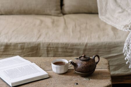 Teapot, cup with beverage, paper book and orchid flower on wood textured table near comfort couch and white wall. Cozy house with wooden interior designの写真素材