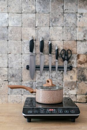 Vertical photo of kitchenware pan at small electric stove with control panel standing in modern kitchen on wooden surface table. Wall with marble stone tile and set of knives on blurred backgroundの写真素材