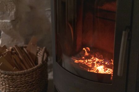 Selective focus on burning coals in wood stove fireplace with metal body and glass door in comfort house with cozy interior in room. Wicker basket with firewood near chimney fireの写真素材