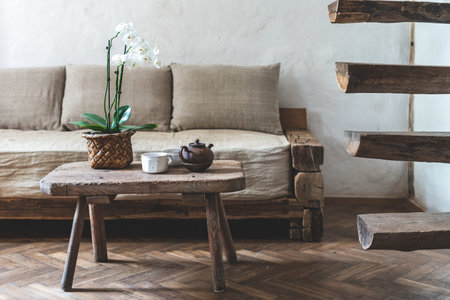 Cozy house with wooden interior design. Teapot, cup with beverage and orchid flower on wood textured table, standing near comfort couch, white wall and retro stairs in room with laminate floorの写真素材
