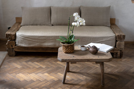 Teapot, cup with beverage, paper book and orchid flower on wood textured table near comfort couch and white wall. Cozy house with wooden interior design in room with laminate floorの写真素材