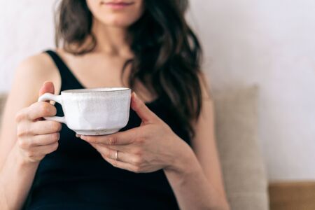 Selective focus of young adult woman holding white cup of beverage in hand. Girl with calm cheerful smile sitting at home near wall with copy spaceの写真素材
