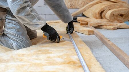 Cropped view of professional workman in protective workwear using knife and holding measuring tape over material, installing thermal insulation rock wool under the roofの写真素材