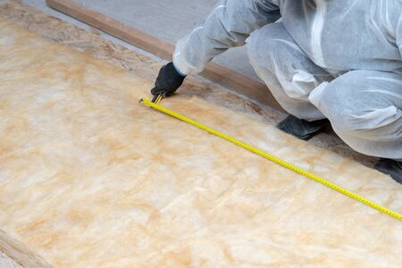 Cropped view of professional roofer man in protective uniform installing thermal insulation rock wool under the roof, using knife and holding measuring tape over materialの写真素材