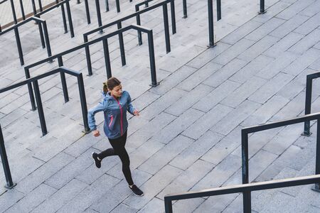 Sporty girl running near stairs against urban background with copy space. High angle view of beautiful and confident young adult athletic woman in sportswear and sneakers training outdoor in cityの写真素材