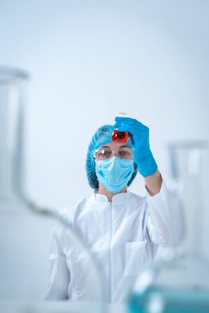 Vertical photo of chemist woman in coat and protective gloves making laboratory experiment, holding medical glass bottles with red liquid, standing isolated on white backgroundの写真素材