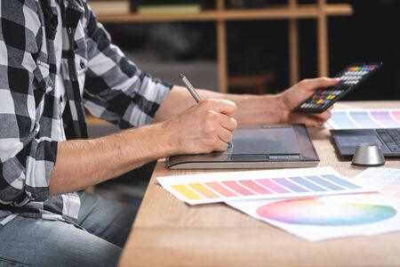 Cropped view of young adult freelancer using color swatches and graphic tablet with stylus, sitting in office, working on computerの写真素材