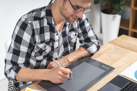 Happy young adult worker smiling wide, using graphic tablet and stylus pencil, sitting and working...