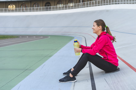 Side view of happy and smiling young adult athlete woman sitting with bottle of water outdoor in...