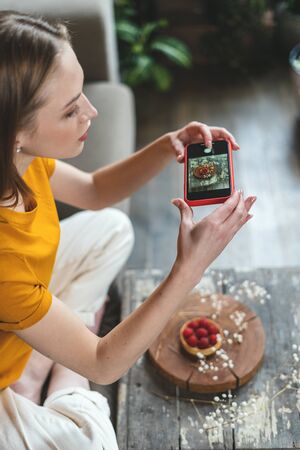 Side view of young adult woman sitting at home, using modern smartphone, making food photo for blog with cupcake dessertの写真素材