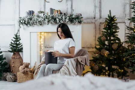 Relaxed young adult girl spending christmas holidays at home, holding present in hands and enjoying seasonal winter traditions in lounge room with decorated new year treeの写真素材