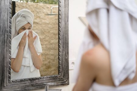 Back view of young adult woman standing in bathroom, looking at mirror reflection, wiping face with white towelの写真素材