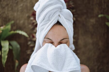 Portrait view of young adult woman with closed eyes standing in bathroom, wiping face with white towel after refreshing, morning routine procedureの写真素材